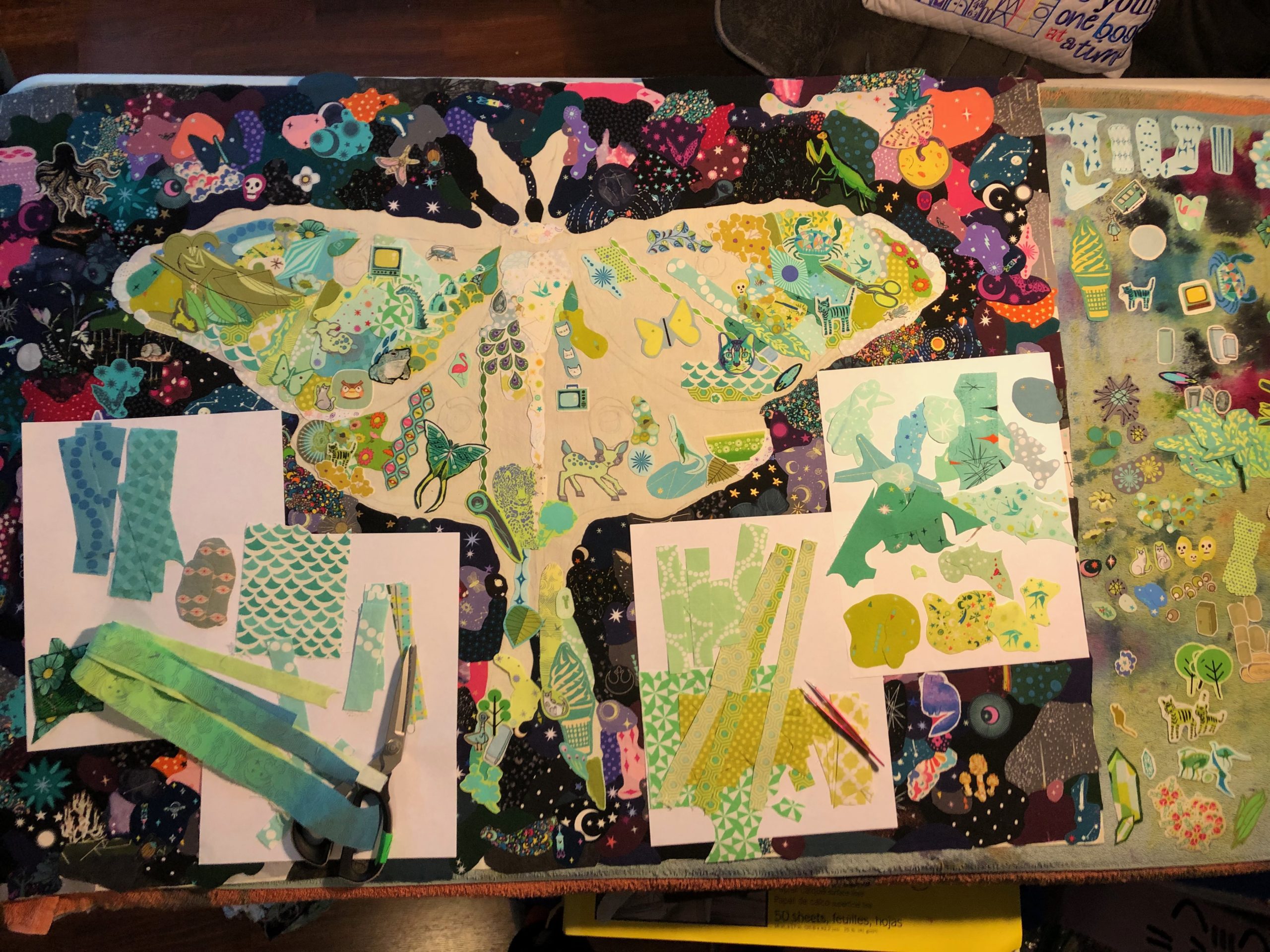 an in-progress collage quilt. the image is of a luna moth. the moth is partially filled in with small pieces of fabric. the background is filled in with small pieces of darker fabrics. some of the fabrics for the moth are on paper on top of the background.
